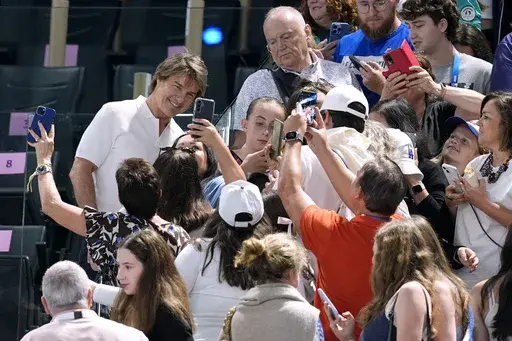 Actor Tom Cruise, left in white, poses for pictures with fans as he attends the women's artistic gymnastics qualification round at the 2024 Summer Olympics, Sunday, July 28, 2024, in Paris, France. (AP Photo/Abbie Parr)