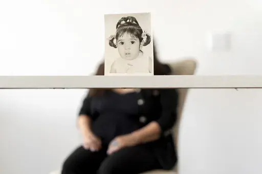 Buttons, who the Associated Press is referring to only by her childhood nickname because of her legal status, sits for a portrait behind her baby photo taken before she was adopted from Iran to a family in America, Monday, June 24, 2024, in Henderson, Nev. Buttons is one of thousands of children adopted from abroad by American parents, many of them military service members, who were left without citizenship by loopholes in American law that Congress has been aware of for decades, yet remains unw