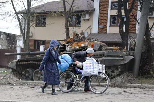 Local civilians walk past a tank destroyed during heavy fighting in an area controlled by Russian-backed separatist forces in Mariupol, Ukraine, Tuesday, April 19, 2022. Taking Mariupol would deprive Ukraine of a vital port and complete a land bridge between Russia and the Crimean Peninsula, seized from Ukraine from 2014. (AP Photo/Alexei Alexandrov)