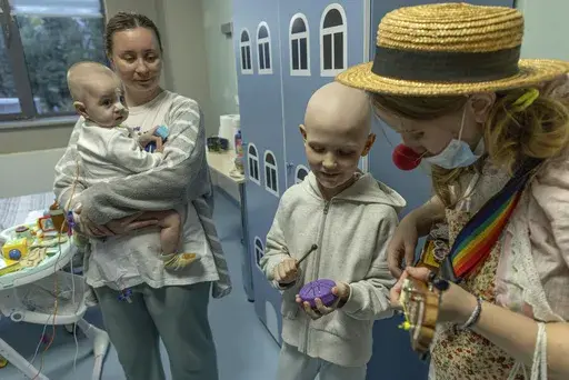 Tetiana Nosova, who goes by the clown name of Zhuzha, a volunteer from the "Bureau of Smiles and Support" plays a ukulele as she stands with Michael Bilyk, who is held by his mother Antonina Malyshko, and Kira Vertetska, 8, at Okhmatdyt children's hospital in Kyiv, Ukraine, Thursday Sept. 19, 2024. (AP Photo/Anton Shtuka)