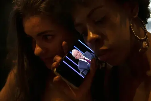 Women listen on a mobile phone to an interview of former president Luiz Inacio Lula da Silva, who is running again for president, with Jornal Nacional on TV Globo, outside a bar in Rio de Janeiro, Brazil, Thursday, August 25, 2022. The Superior Electoral Court, the country's top electoral authority, announced Thursday, Oct, 20, that it would be banning "false or seriously decontextualized" content that “affects the integrity of the electoral process.” No request from a prosecutor or complain