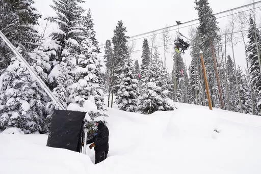Ian Sidwell adjusts a machine used to make snow at Vail Mountain Resort as snowboarders ride a lift, Wednesday, Dec. 29, 2021, in Vail, Colo. Newer snowmaking technology is allowing ski areas to be more efficient with energy and water usage as climate change continues to threaten snowpack levels. (AP Photo/Brittany Peterson)